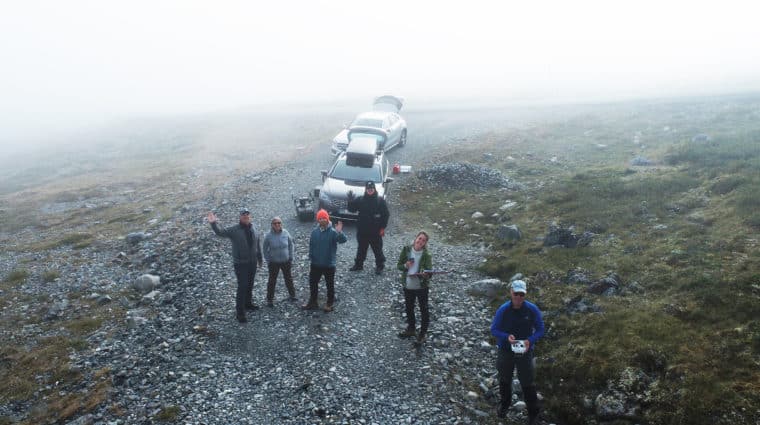 Taken from above, six people are waving at the camera. Two cars sit behind the people, who stand on a road with fog behind them.