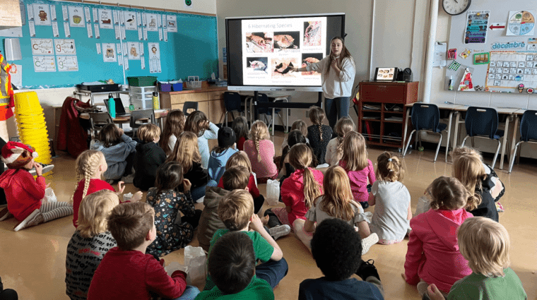 A person standing at a screen talking to a classroom of children who are sitting on the floor.