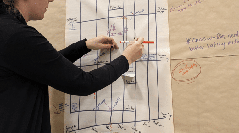 A person stands at a board looking at a paper with a grid like drawing. The person is holding a red marker.