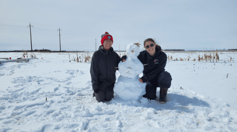 Two people in a field sitting next to small snowman.